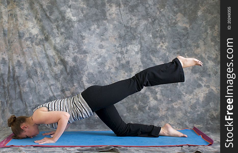 Young woman on yoga mat in Yoga posture sunbird pose with left leg lifted, against a grey background in profile, facing left lit by diffused sunlight. Young woman on yoga mat in Yoga posture sunbird pose with left leg lifted, against a grey background in profile, facing left lit by diffused sunlight.