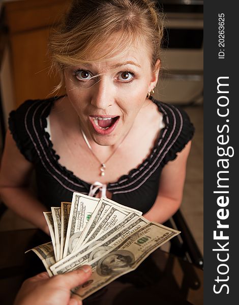 Blond woman in casual attire with a shocked expression sitting at home with hand giving lots of 100 dollar bills. Blond woman in casual attire with a shocked expression sitting at home with hand giving lots of 100 dollar bills