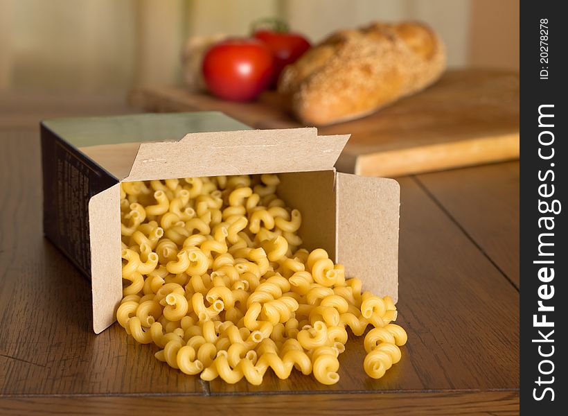 Uncooked corkscrew shaped pasta spilling from the box, with a loaf of Italian bread and tomatoes in the background. Uncooked corkscrew shaped pasta spilling from the box, with a loaf of Italian bread and tomatoes in the background