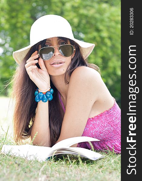 An attractive woman wearing a hat and sunglasses reading a book on the park looking at the camera. An attractive woman wearing a hat and sunglasses reading a book on the park looking at the camera