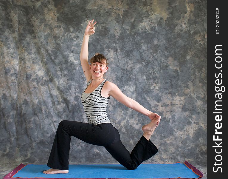 Young woman on yoga mat doing Yoga posture King Arthur's pose variation, with arm raised and hand in om position, looking at viewer and smiling, against a grey background in profile, facing left lit by diffused sunlight. Young woman on yoga mat doing Yoga posture King Arthur's pose variation, with arm raised and hand in om position, looking at viewer and smiling, against a grey background in profile, facing left lit by diffused sunlight.