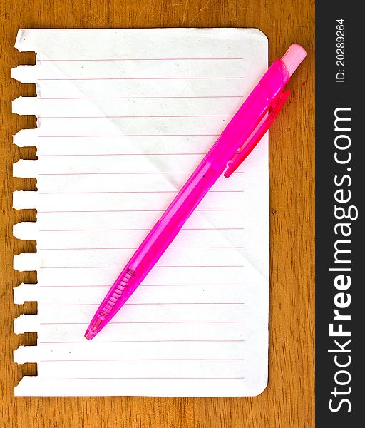 The pink pen on the empty notebook paper and on the wood. The pink pen on the empty notebook paper and on the wood