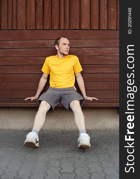 Man in yellow shirt sitting on bench near wooden wall, looking at side