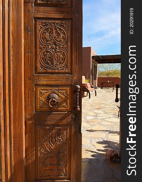 A wooden, ornate, opened door with metal knocker leads to a inner courtyard with stone and seating. A wooden, ornate, opened door with metal knocker leads to a inner courtyard with stone and seating