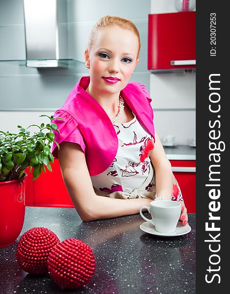 Young beautiful blond girl wearing white and pink dress with cup of morning coffee in interior of red modern kitchen. Young beautiful blond girl wearing white and pink dress with cup of morning coffee in interior of red modern kitchen