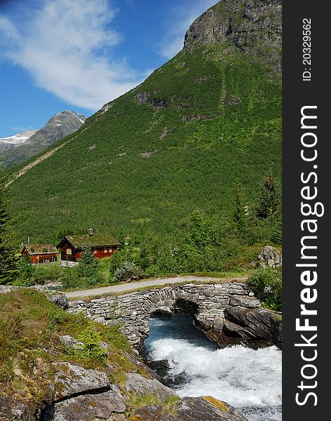 Small wooden houses with greenroof are located under the green mountain. Rapid river is running under stone bridge in the foreground. Small wooden houses with greenroof are located under the green mountain. Rapid river is running under stone bridge in the foreground.