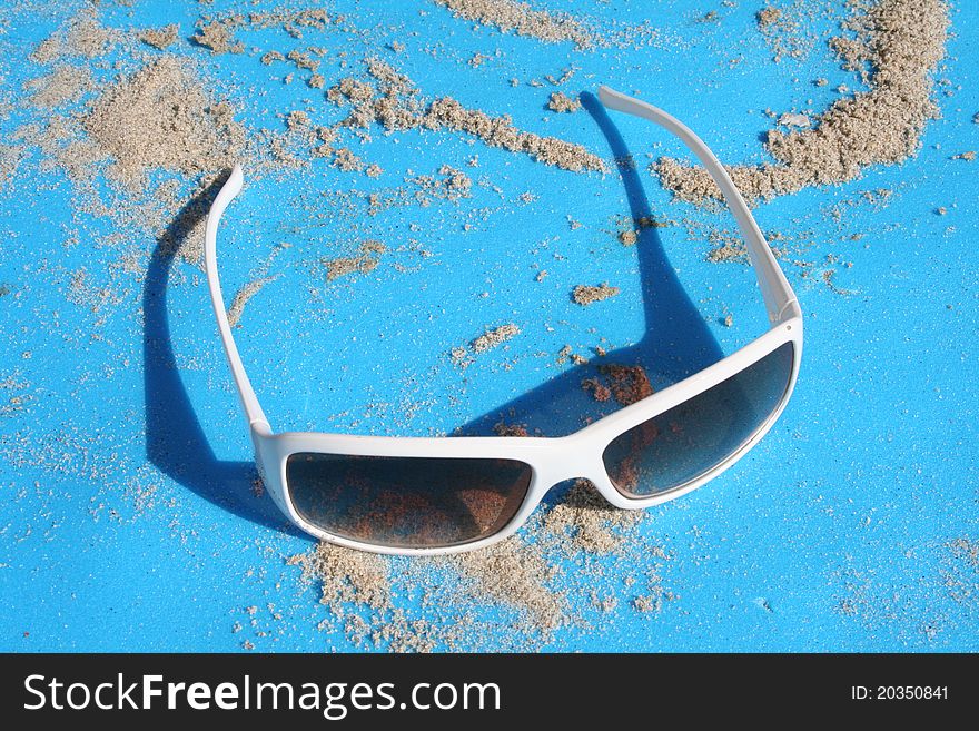 White sunglasses sitting on a blue backround with sand.