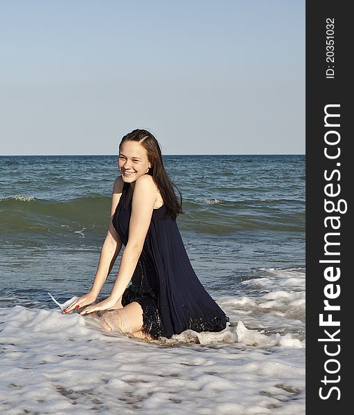 Beautiful young girl in black wet dress sits in water of the sea