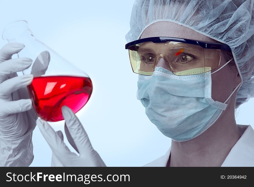 Female, caucasian lab technician with goggles and protective gear studying conical flask with red liquid. Female, caucasian lab technician with goggles and protective gear studying conical flask with red liquid