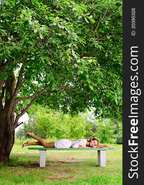 Pretty barefooted woman with is laying on a bench under the green tree. She is slightly smiling and looking at the camera. Pretty barefooted woman with is laying on a bench under the green tree. She is slightly smiling and looking at the camera