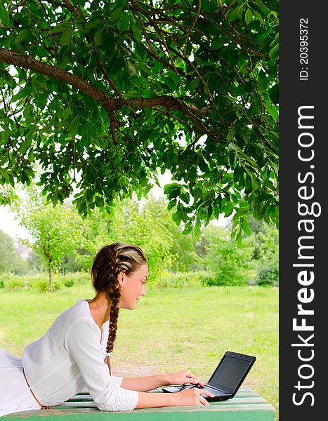 Pretty barefooted woman is laying on a bench under the green tree. She is working with small notebook. Pretty barefooted woman is laying on a bench under the green tree. She is working with small notebook