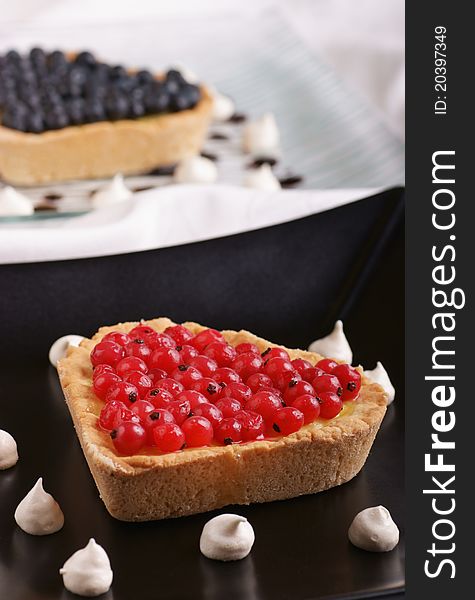 Heart-shaped tart with redcurrant served on a black plate with small meringues. A tart with blackcurrant is out of focus in the background
