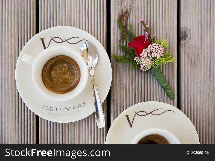 Couple of cups with Italian Espresso, a teaspoon and flowers a wedding boutonniere, on wooden table. Vistamare means: seaview. Couple of cups with Italian Espresso, a teaspoon and flowers a wedding boutonniere, on wooden table. Vistamare means: seaview