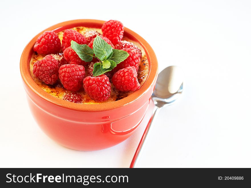French creme brulee dessert with raspberries and mint covered with caramelized sugar in red terracotta ramekin on white background