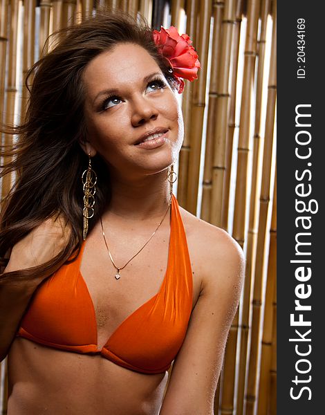 Beautiful sexy girl with a red flower in long hair in an orange bikini on the sunny beach. She is isolated on a bamboo background. Beautiful sexy girl with a red flower in long hair in an orange bikini on the sunny beach. She is isolated on a bamboo background