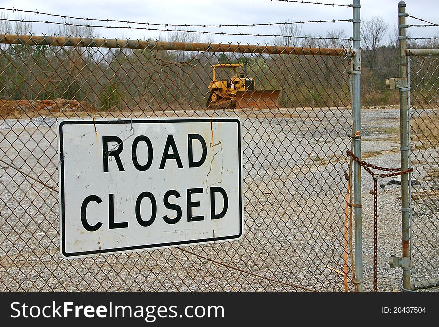 A road closed sign fastened to a chain link fence, a bulldozer in the background