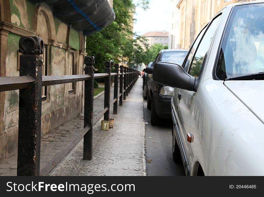 Parked cars in the city. Fence is between the road and the sidewalk. Old houses near to the sidewalk. Parked cars in the city. Fence is between the road and the sidewalk. Old houses near to the sidewalk.