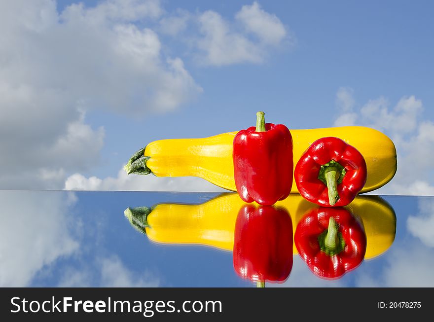Two red peppers and courgette on mirror and sky