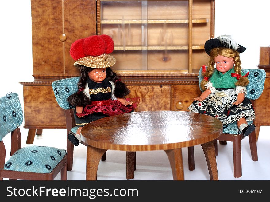 2 dolls in traditional European (I think they are Austrian but not sure) dresses sitting on chairs round a table (old hand made furniture) with racks on the background. 2 dolls in traditional European (I think they are Austrian but not sure) dresses sitting on chairs round a table (old hand made furniture) with racks on the background.