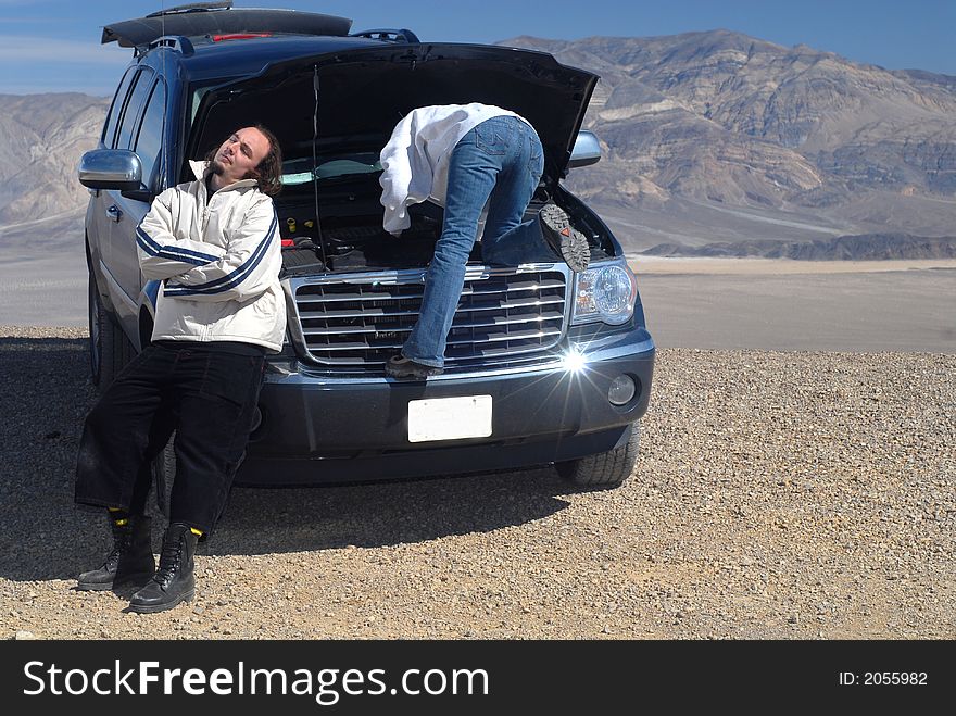 Woman Trying To Fix Car While Husband Relaxes