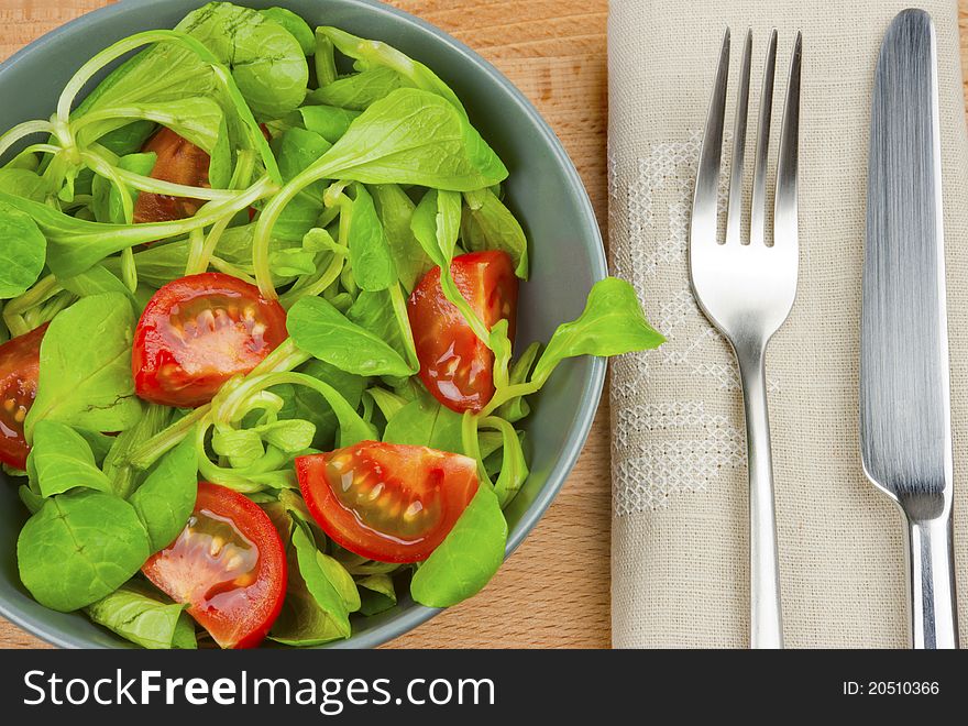 A salad of lettuce and tomatoes in a bowl, metal knife and fork on napkin linen on a wooden table. A salad of lettuce and tomatoes in a bowl, metal knife and fork on napkin linen on a wooden table