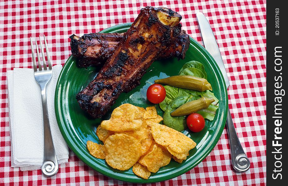 Bar-B-Que Ribs on a checkered table cloth with silverware, napkin, salad and chips.