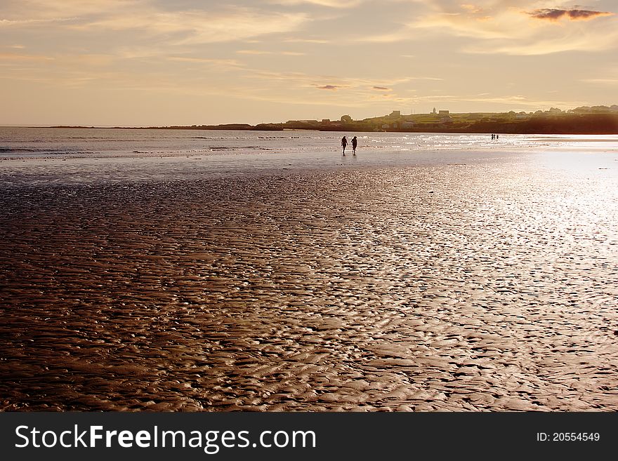 Two people walk along the shore of a quite beach at low tide. Two people walk along the shore of a quite beach at low tide