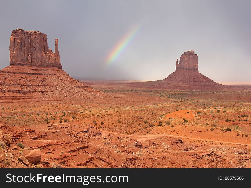 Rainbow forming between Mitten rock formations in Monument Valley, Arizona with winding road in foreground. Rainbow forming between Mitten rock formations in Monument Valley, Arizona with winding road in foreground