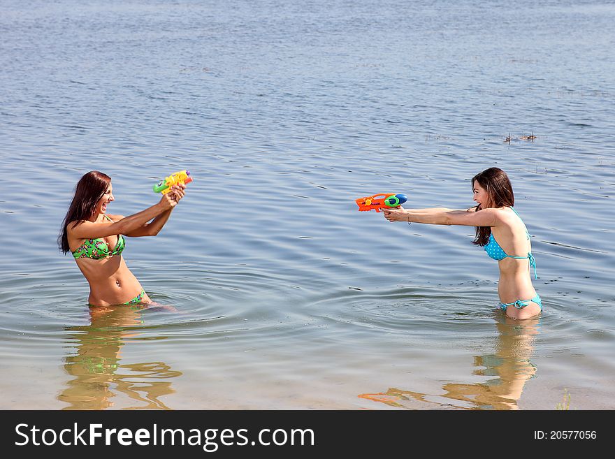Two attractive girls in the swimsuits on the beach play with a water guns. Two attractive girls in the swimsuits on the beach play with a water guns