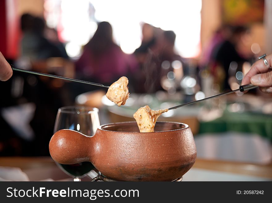 Two people serving fondeau from a ceramic hot bowl in a restaurant and a cup of wine behind, with other people having a meal in the blury background.