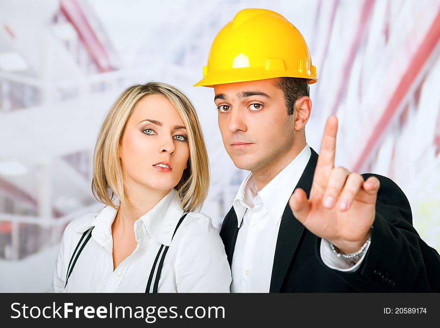 Male and female architects looking at camera, man wearing hardhat and pointing with finger. Male and female architects looking at camera, man wearing hardhat and pointing with finger