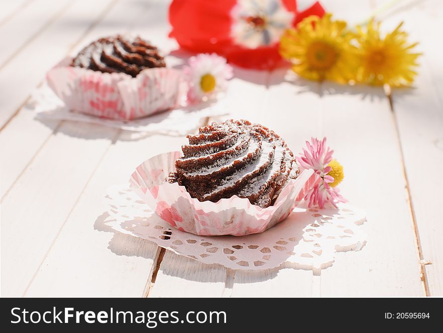 Two chocolate cakes in paper baking cups on the table with flowers. Two chocolate cakes in paper baking cups on the table with flowers