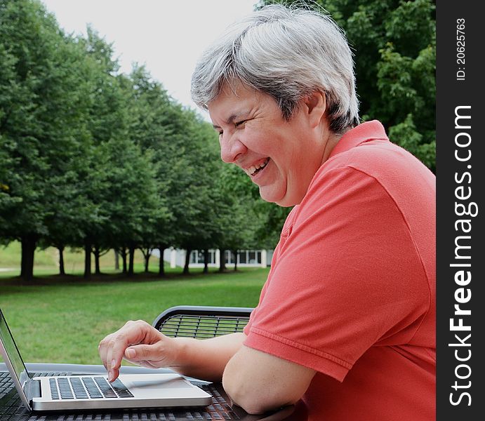 Middle-aged woman browsing on laptop at table outside with trees. Middle-aged woman browsing on laptop at table outside with trees