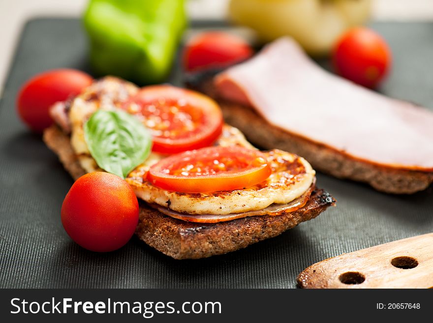A cheese, ham, tomato and basil sandwich with whole rye bread on a cutting board with wooden utensil. A cheese, ham, tomato and basil sandwich with whole rye bread on a cutting board with wooden utensil.