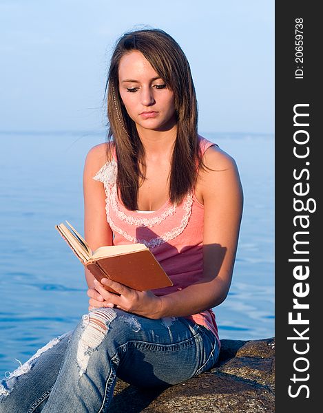 Woman reading on a rock at the edge of an ocean or seaside or body of water.
