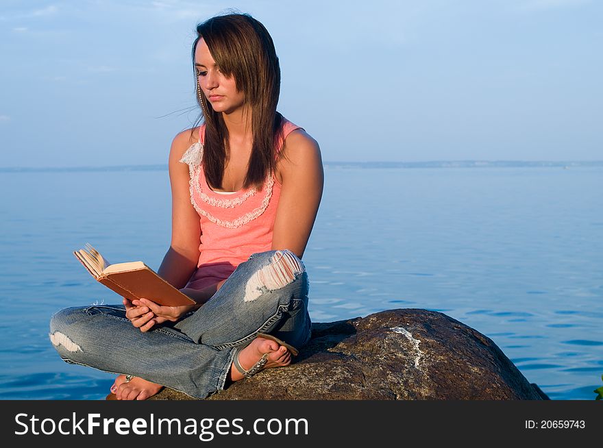 Woman reading on a rock at the edge of an ocean or seaside or body of water. She is at peace and enjoying her downtime.