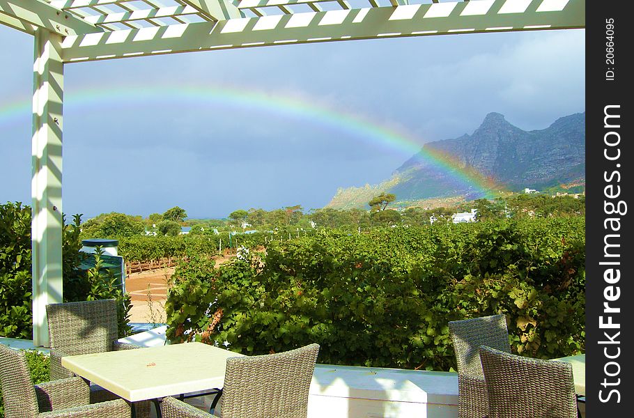 A sitting area foreground with view over a vineyard and mountains. A rainbow emerges after a light rain. A sitting area foreground with view over a vineyard and mountains. A rainbow emerges after a light rain