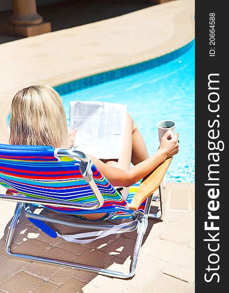 The woman is holding cup of coffee and reading newspaper by the pool. The woman is holding cup of coffee and reading newspaper by the pool