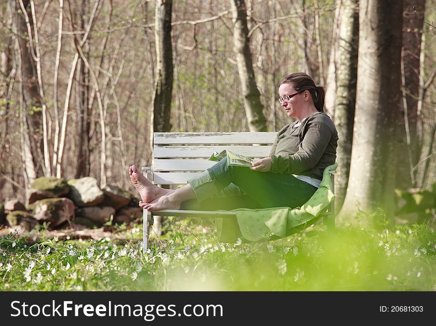 Woman sitting on a sofa in the wood in springtime. She is reading a magazine in the sunlight. Woman sitting on a sofa in the wood in springtime. She is reading a magazine in the sunlight