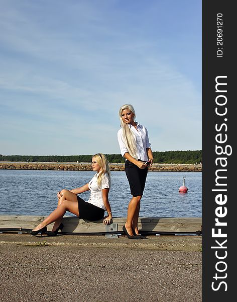 Two blonde girls on the beach. One is standing and looking at the camera. Another sits. Two blonde girls on the beach. One is standing and looking at the camera. Another sits.