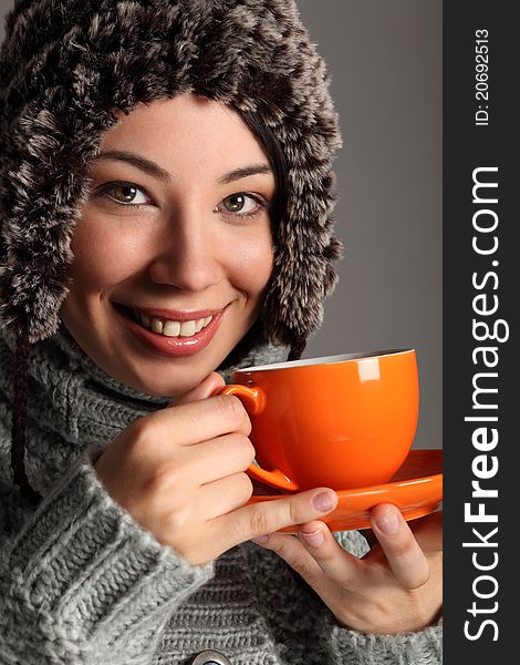 Beautiful smile from young woman wearing thick wool sweater and furry winter hat, drinking tea from orange cup. Shot against a grey background. Beautiful smile from young woman wearing thick wool sweater and furry winter hat, drinking tea from orange cup. Shot against a grey background.