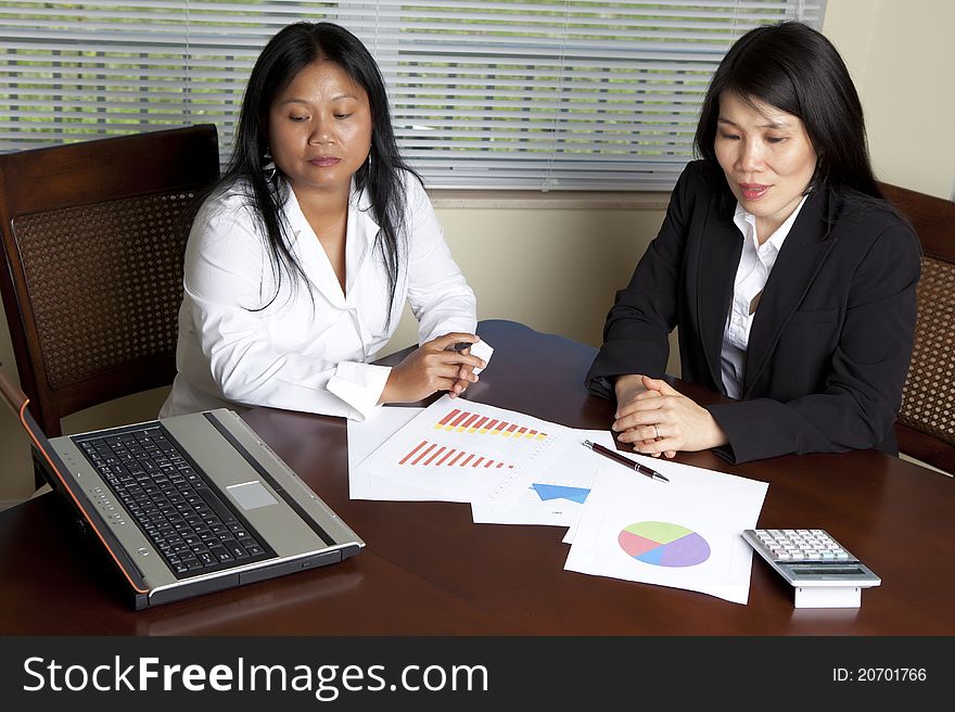 Two Asian Women at desk with laptop charts, graphs on desk.