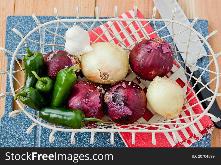 Jalapeno peppers, red onions, yellow onions, and a head of garlic rest in a wire basket on top of a cutting board next to a chef knife and red stripe towel. Jalapeno peppers, red onions, yellow onions, and a head of garlic rest in a wire basket on top of a cutting board next to a chef knife and red stripe towel.