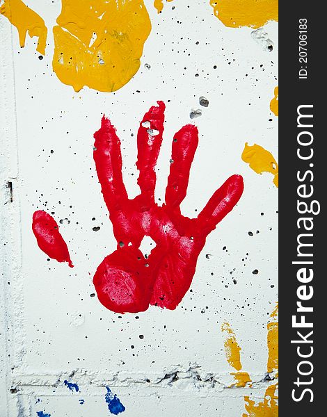 A child's handprint in red paint on a white stone wall. Other paint spots in yellow and blue are also seen.