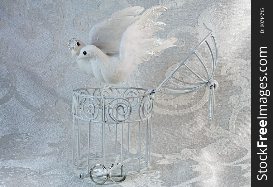 Doves resting on an open white painted cage with wedding rings on a brocade textile. Doves resting on an open white painted cage with wedding rings on a brocade textile