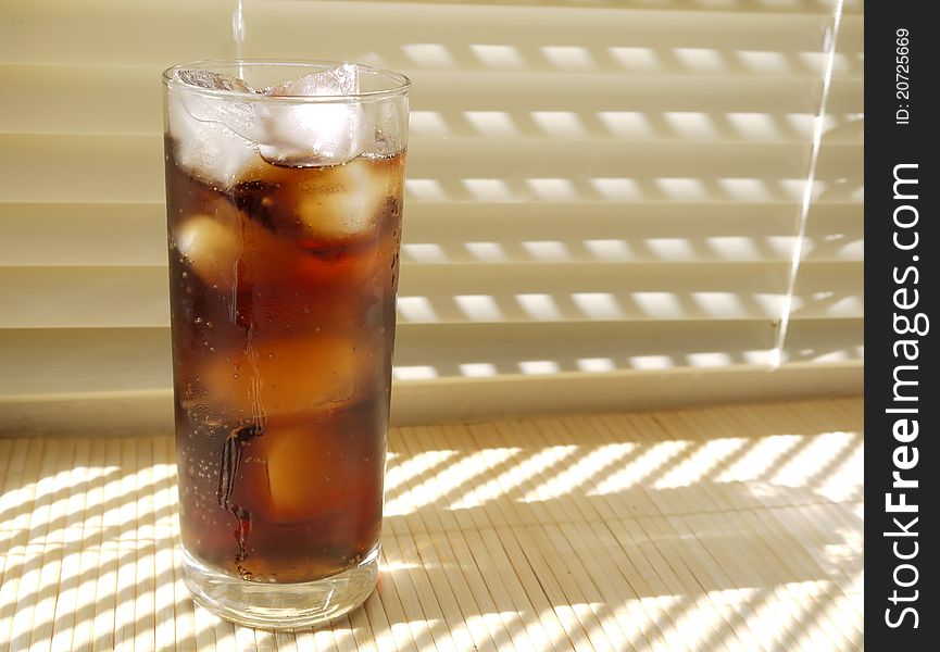 An ice cold fizzy drink with ice in a tall glass on a cream blind background on a sunny day. An ice cold fizzy drink with ice in a tall glass on a cream blind background on a sunny day