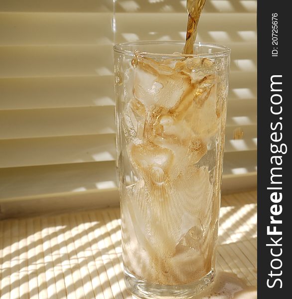 A Tall glass of ice with fizzy cola pouring into it on a cream blind background on a sunny day. A Tall glass of ice with fizzy cola pouring into it on a cream blind background on a sunny day