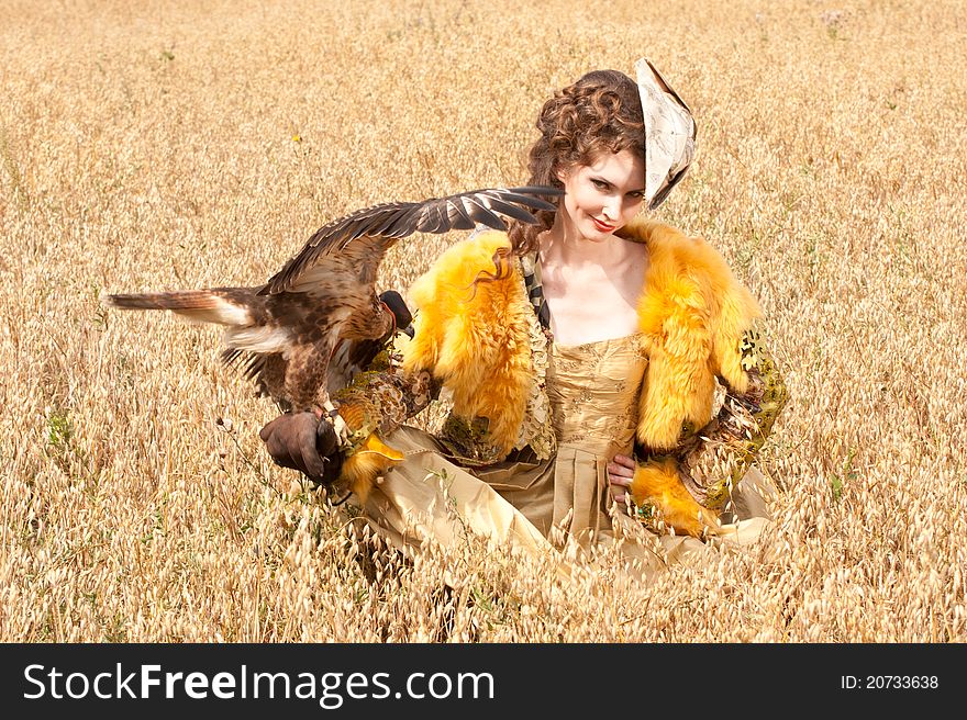 The woman in a beautiful old style dress with falcon has a rest before hunting in yellow field. The woman in a beautiful old style dress with falcon has a rest before hunting in yellow field.
