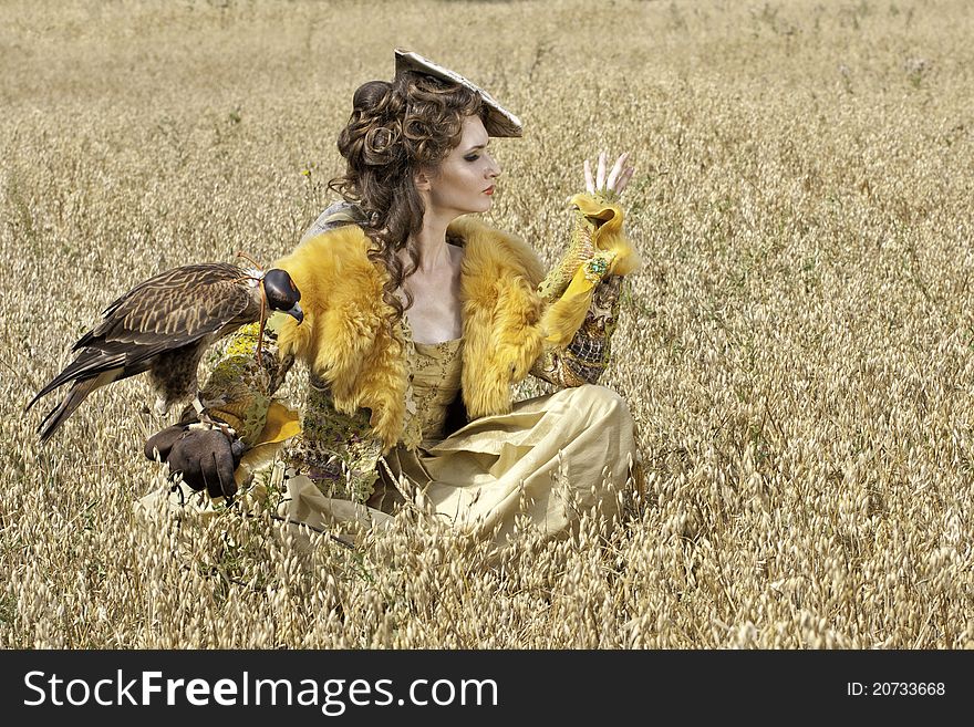 The woman in a beautiful old style dress with falcon has a rest before hunting in yellow field. The woman in a beautiful old style dress with falcon has a rest before hunting in yellow field.