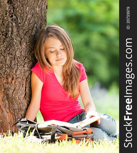 Beautiful and happy young student girl sitting on green grass under the tree, smiling and reading book. Summer or spring green park in background. Beautiful and happy young student girl sitting on green grass under the tree, smiling and reading book. Summer or spring green park in background